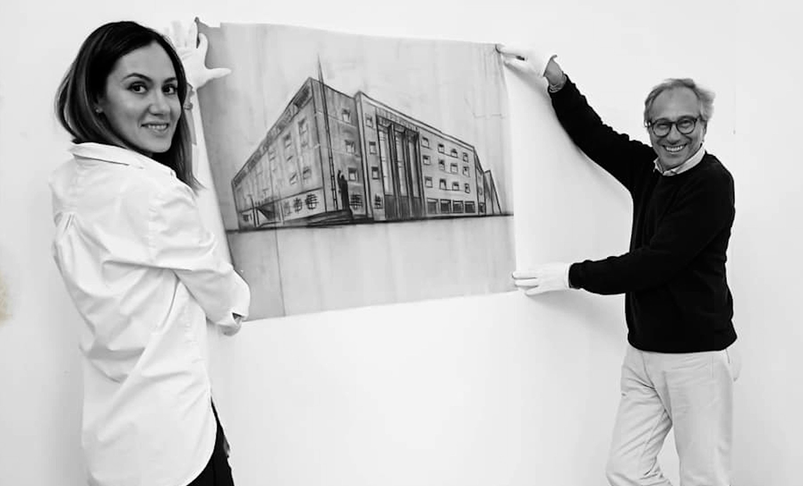 Black and white photograph of two people holding a large architectural drawing of a multistory building against a plain wall