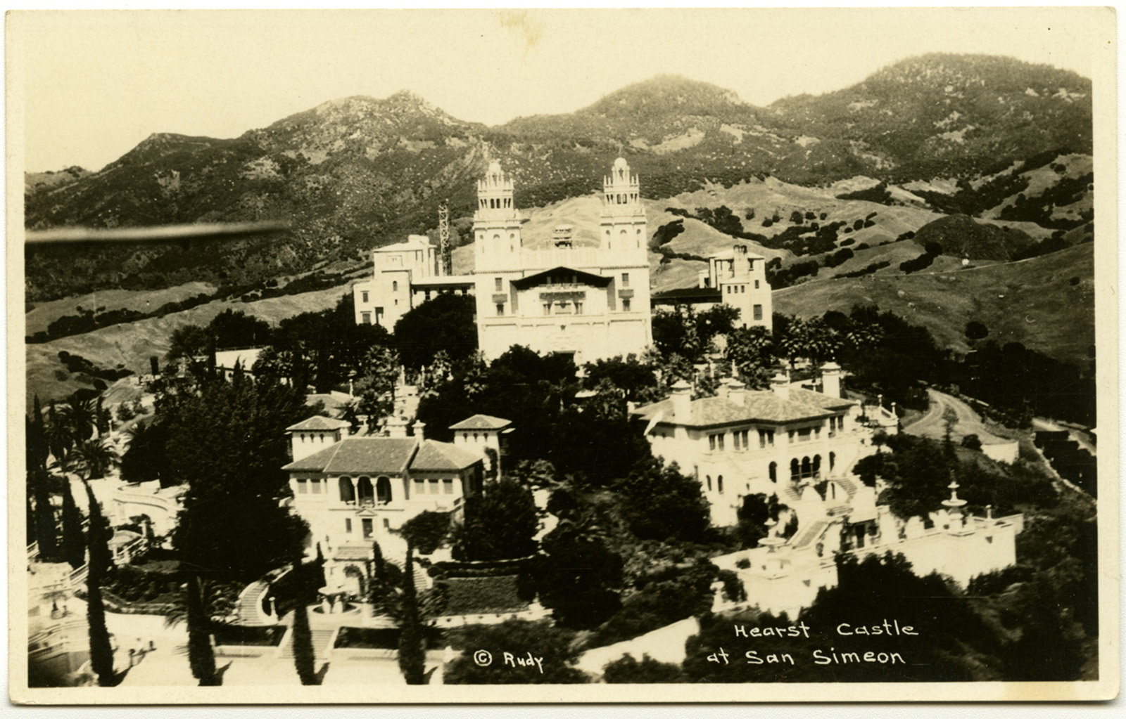 A vintage photograph of Hearst Castle at San Simeon, set on a hill with surrounding greenery and mountains. The castle is prominently featured with its distinct towers and intricate architecture.