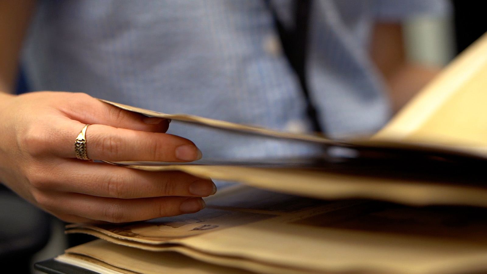 A person wearing a ring gently flips through the pages of an old book or document. The focus is on the hand and the pages, suggesting a task that requires attention and care.