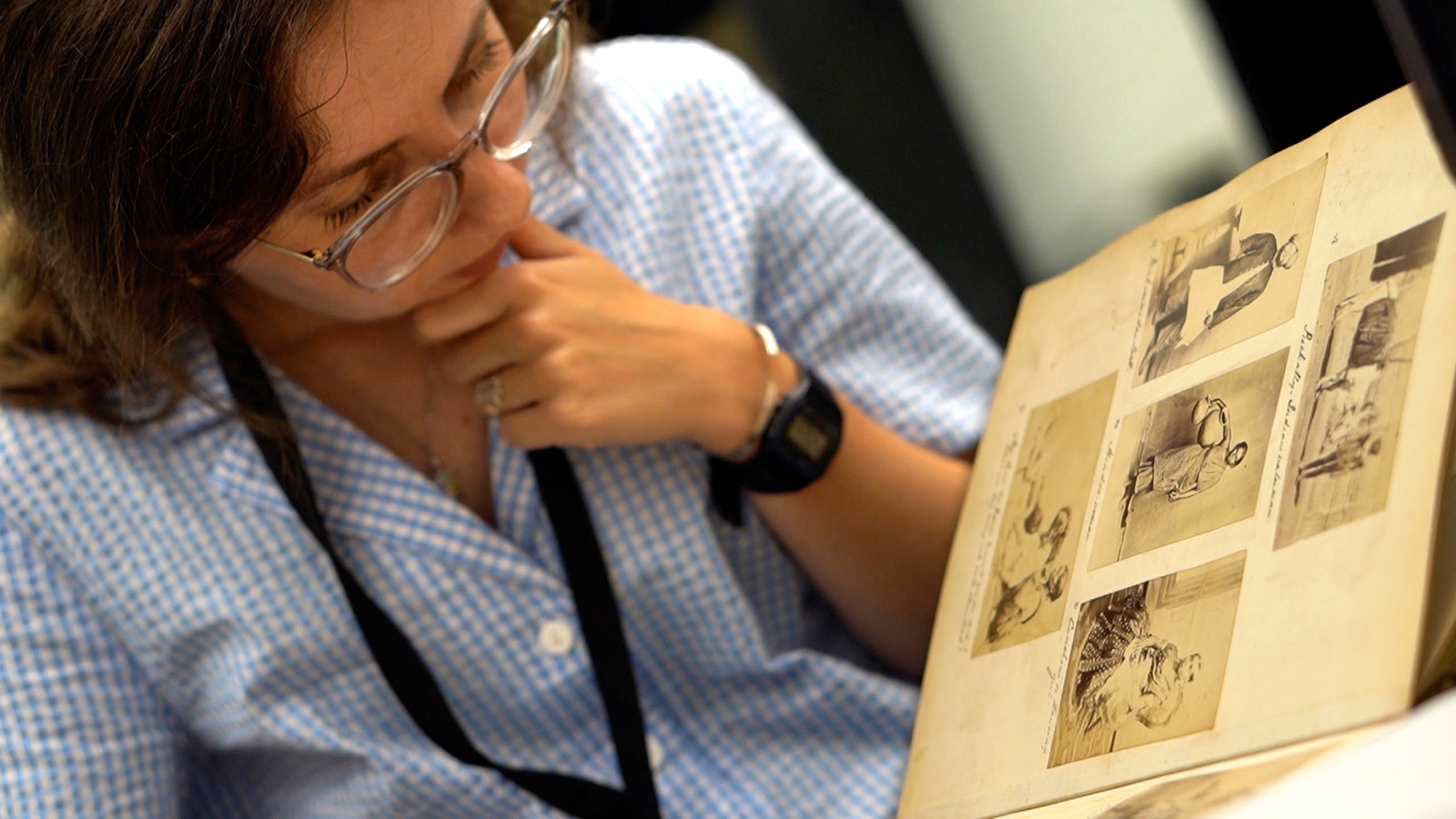 A person wearing glasses examines a photograph album, dressed in a blue checkered shirt and wearing a lanyard.