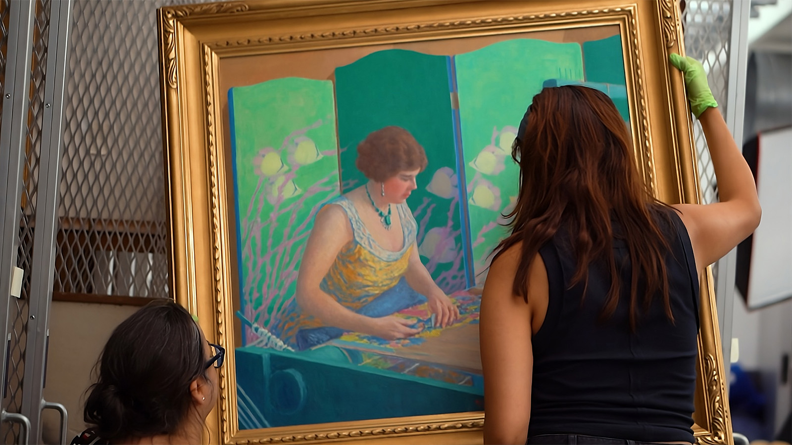 Two women handle a large framed painting of a seated woman at a table, positioning it upright in front of a metal hanging rack.