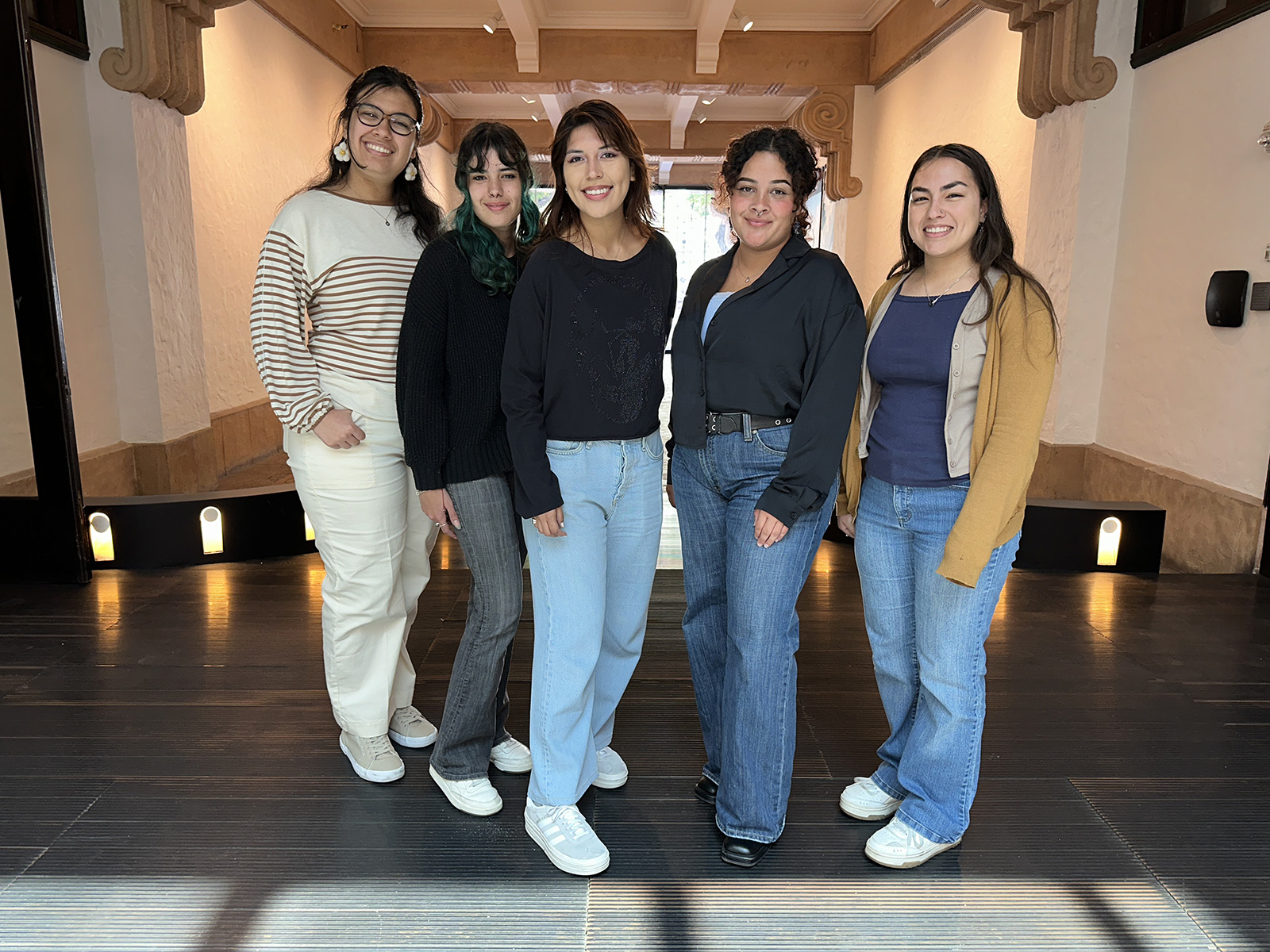 Five young women stand together in a lit hallway, smiling at the camera, dressed in casual sweaters and jeans.