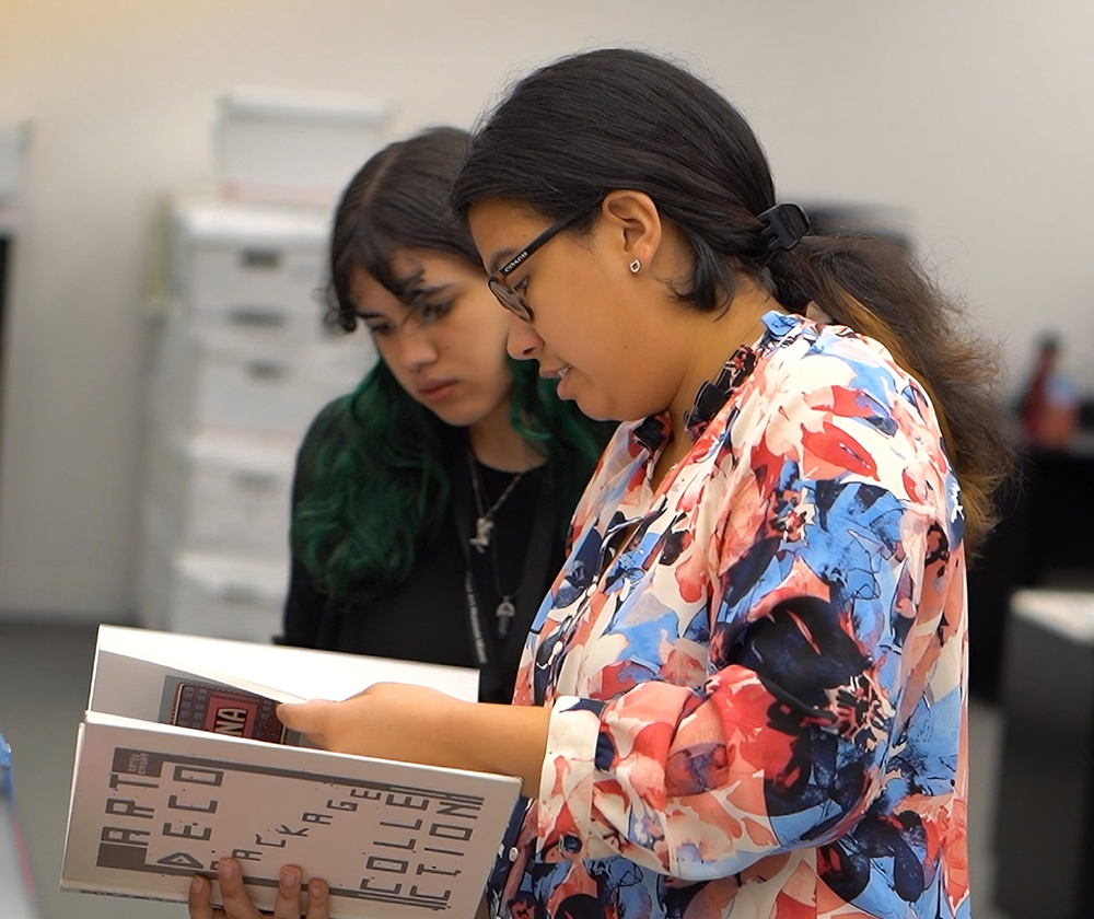 Two women look at an open book together in a workspace lined with shelves and storage boxes.