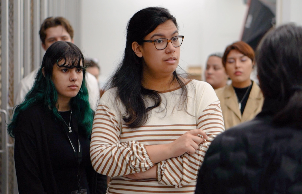 A group of students stands together indoors, with two women in the foreground facing a person who is speaking in partial view of the camera..