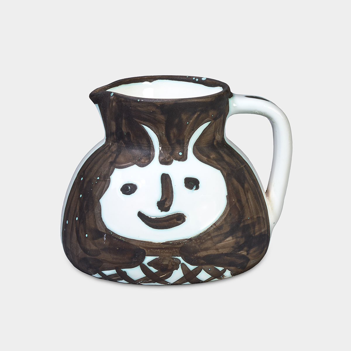 A smiling face appears on the surface of this ceramic pitcher. The side facing us is painted dark brown, while the area around the face is left unpainted, revealing the stark white color of the ceramic. The face is large and rounded, featuring wide, oval eyes, a dark brown line for a nose, and a curved mouth. Two horns protrude from the top of the head. Below the face, the neck and shoulders are white and centered at the base of the pitcher, adorned with thick, crossed dark brown lines.  To our right, the handle of the pitcher has spots of dark brown paint along its length but remains mostly white. The mouth and rim at the top of the pitcher are painted dark brown, while the interior retains the same white color as the ceramic.