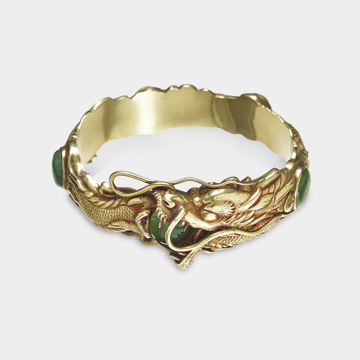 A gold cuff cast in the shape of a dragon contains five evenly-spaced jade gemstones embedded into the metal. The dragon is positioned with its body coiled and head turned to the side, its mouth open revealing sharp teeth and wrapping around one of the jade stones. The dragon’s scales are individually defined, and its eyes are prominent and of a reflective quality. The snout of the dragon has two long whiskers that curl and flow down the back of its head, slightly lifting off of its body. The cuff is primarily metallic, with a shiny, reflective surface in shades of bronze and gold. The inside of the cuff is smooth and unadorned.