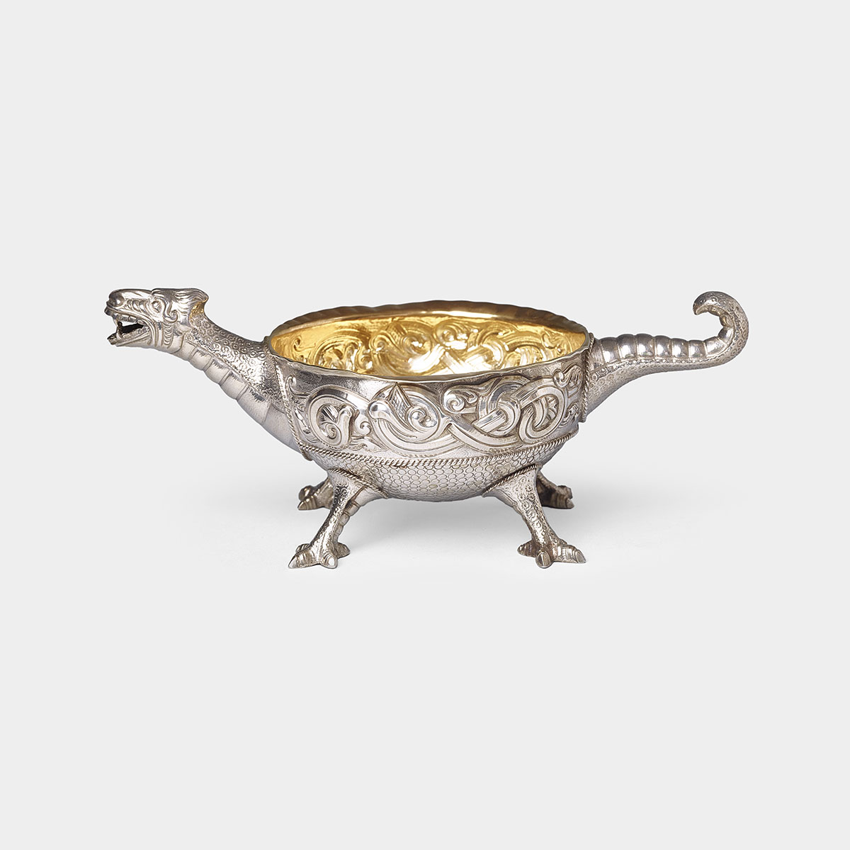 A silver bowl cast in the shape of a dragon contains a gold interior and is supported by four small, outstretched dragon feet. The dragon’s head and neck extend from one side of the bowl, and the dragon’s long, curled tail follows out of the opposite end at a raised angle. The dragon’s mouth is open, revealing sharp teeth, and its eyes are rounded and open. The dragon’s body, which makes up the form of the bowl, is scaly in texture at the bottom and decorated with ridges and swirled patterns along the top. The legs of the dragon, which sit under the bowl’s base, are muscular and end in sharp, clawed feet to support the object. The entire bowl is metallic, with a slightly reflective surface.