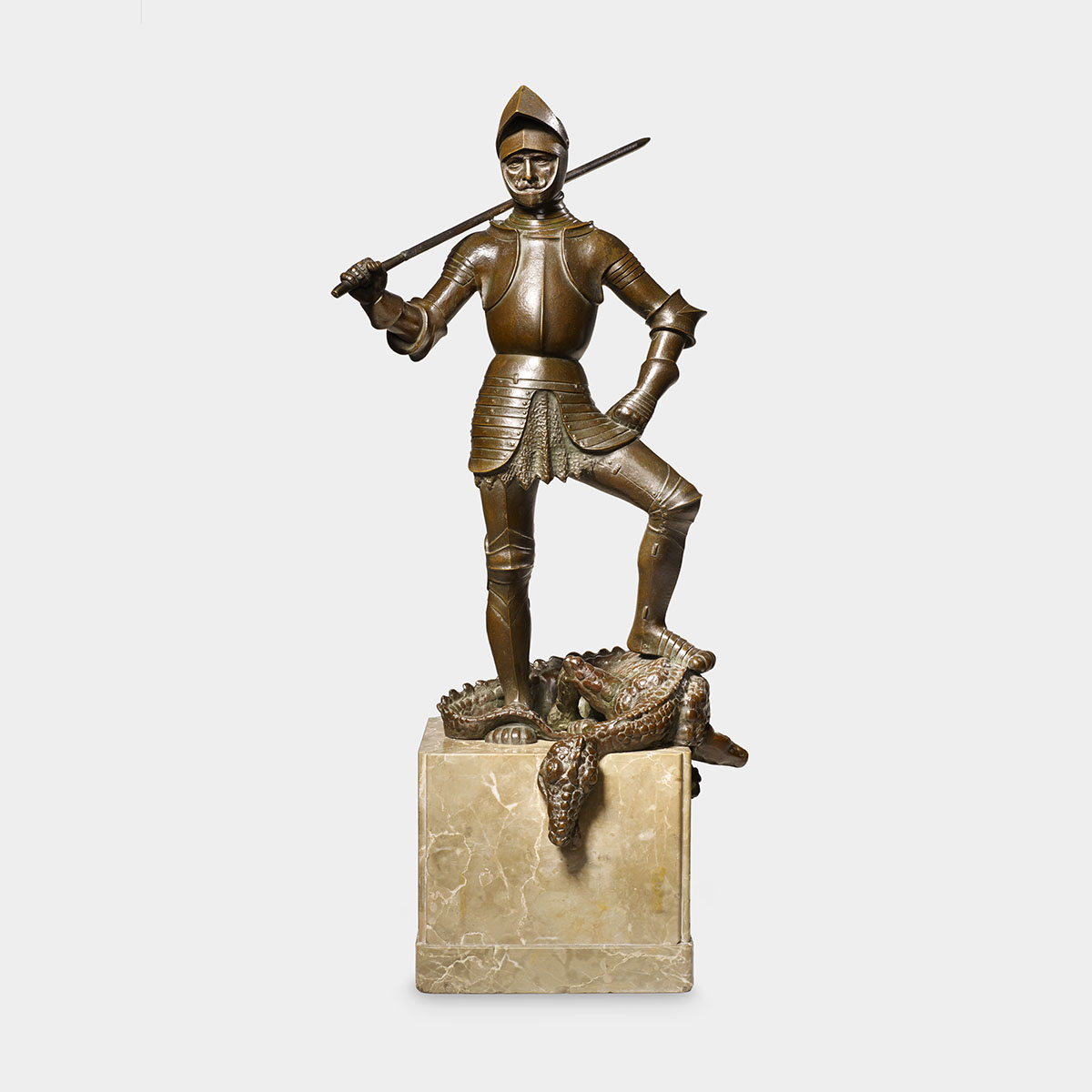 A man dressed in armor carrying a sword that rests on his right shoulder stands over a dragon in this bronze sculpture. He wears a helmet with the face covering lifted, a breastplate, armor for his legs and arms, and chainmail underneath. The man stands with his left foot placed over the dragon’s body and his right foot on the cubed, pillar-like base of the sculpture. His left arm is bent, hand resting on his hip, and his right arm is bent and lifted slightly upward to support his sword. The man’s face is partially visible through the helmet, his eyes narrowed and staring directly at us. The dragon beneath the man has a scaly body with a long, curved tail that drapes over the edges of the sculpture’s base. Its head hangs down over the front of the sculpture, slightly turned to the side and eyes closed. The man and dragon have a brown, bronze color with a glossy finish, and the base is made of a polished stone with a pale, beige tone and visible marble-like veins.