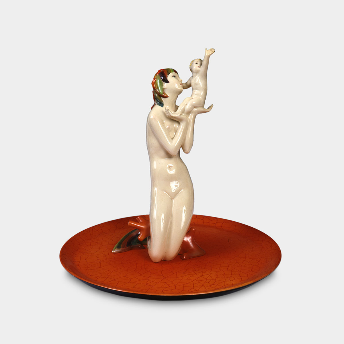 A nude female figure kneels at the center of a bright orange plate, holding a small child in this ceramic sculpture. Both figures have light, pale skin and slender physiques. The woman faces forward, her head tilted upward with wide-open eyes, gazing to the right at the child in her raised hands. She has short, stylized hair streaked with red, green, and black. Her lower legs gradually transform into colorful fins of the same hues, which rest on the orange base. The child, with pale yellow hair, sits in the woman's hands. His right arm is positioned just behind her head, while his left arm stretches upward above his face. His head is tilted back and to the right, with his mouth slightly open, and his body faces left, toward the woman. The orange plate serving as the sculpture's base features a cracked glaze pattern and slightly upturned edges.