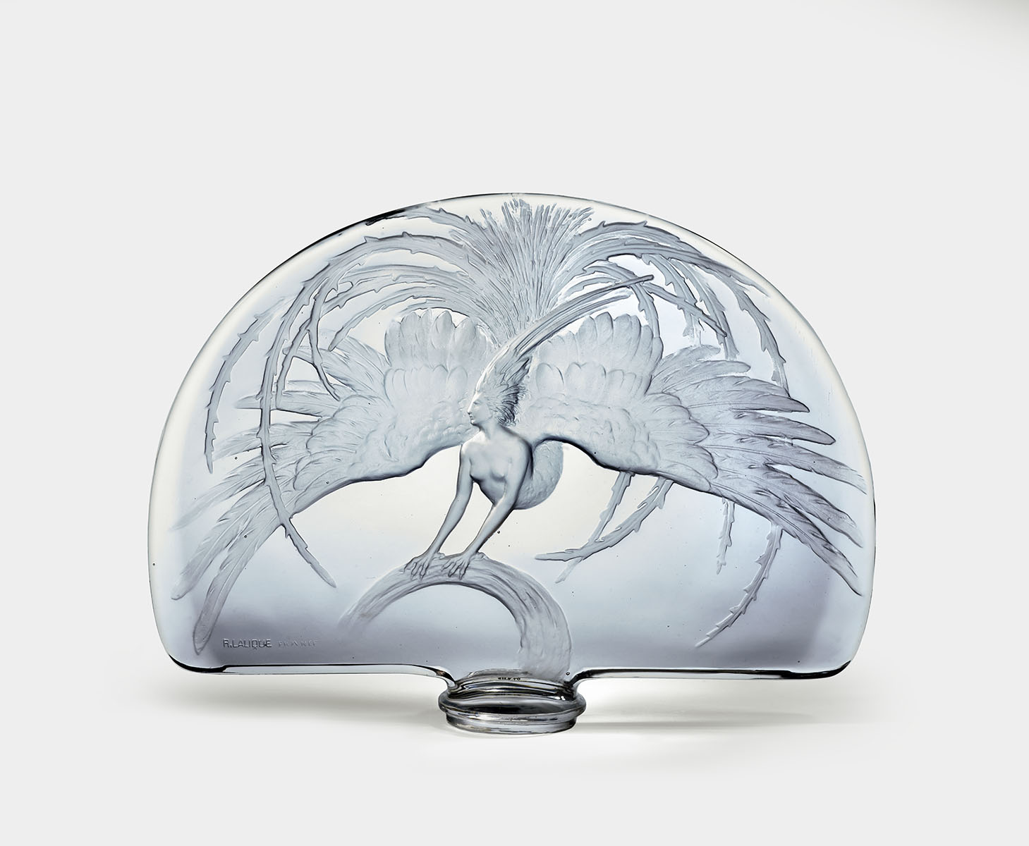 A woman with a human upper body and a bird-like lower body spreads her feathers in the etched design of this glass centerpiece. The glass piece has a semi-circular shape, flat on the bottom with a rounded top, and is mounted on a small, circular base. The woman depicted has large, outstretched bird wings with detailed feather etchings. The wings are spread wide, extending almost to the edges of the glass, and the tail feathers curve downwards, following the shape of the sculpture. The woman’s face is seen in profile view and turned to our left, while her nude upper body faces us slightly turned to the left. Her arms are stretched out in front of her, with hands resembling bird talons grasping a curved, branch-like structure drawn along the central bottom edge of the sculpture. Large, curved feathers fan out behind the woman in the background, some bending forward into the foreground. The glass has a smooth, transparent finish, with frosted, slightly opaque details that emphasize the etched portions of the sculpture.