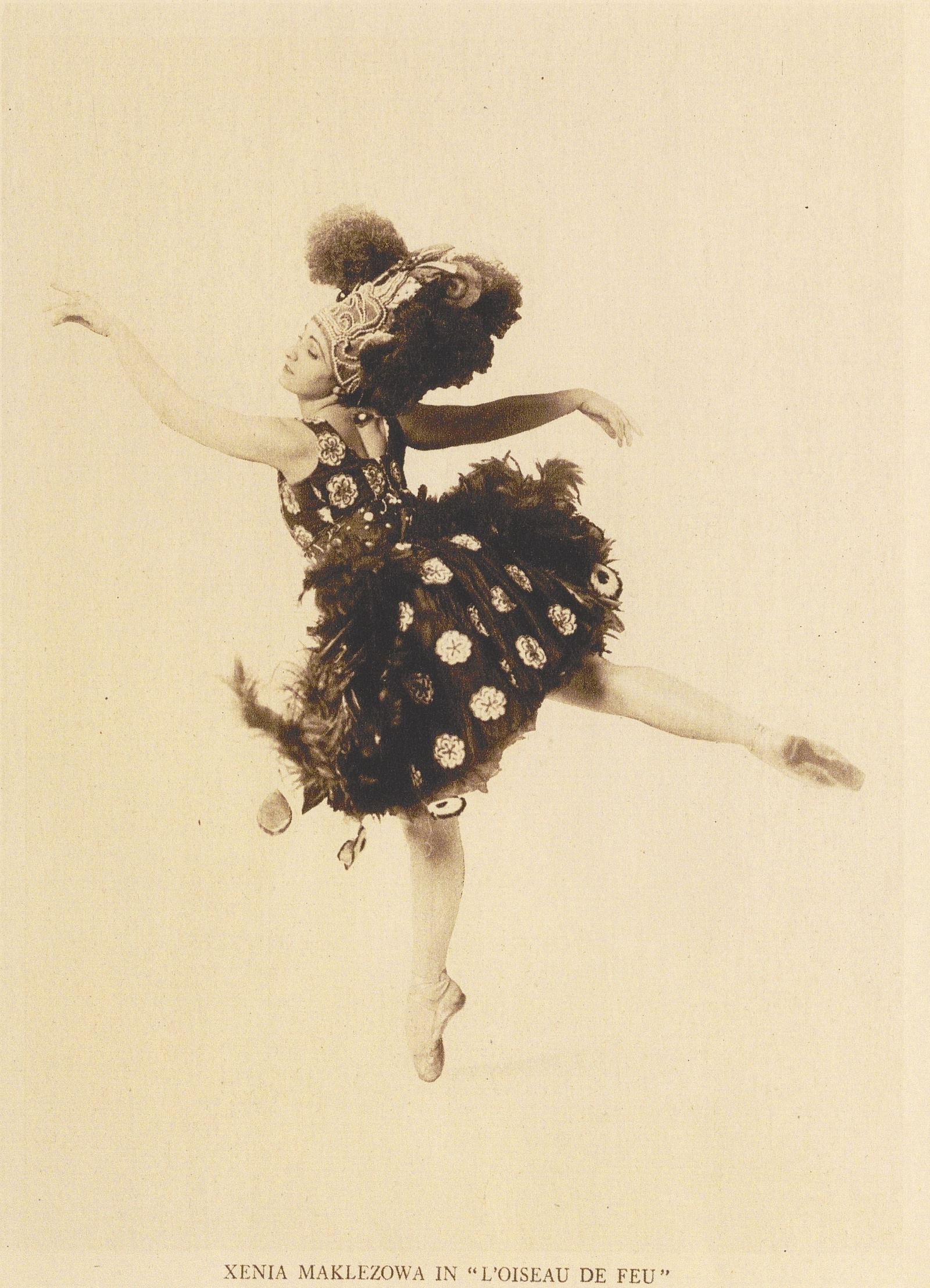 A woman dressed in a bird-like costume dances at the center of this illustration. The dancer, identified by the text at the bottom as "Xenia Maklezowa in 'L'Oiseau de Feu'," is posed with one leg raised behind her and both arms outstretched. She has light skin and is drawn using a sepia tone for her clothing. Her costume consists of a sleeveless bodice and a knee-length skirt, both adorned with circular floral motifs. The skirt appears voluminous with layers and features large feather-like decorations along the edges. The dancer also wears a large, ornate headdress with intricate designs and curved plumes and feathers that extend upward, fanning out behind her head. Her facial expression is serene, with her head slightly tilted back and eyes closed. Her ballet shoes and the lower part of her legs are visible, with her left foot pointed and her right foot raised, capturing the sense of movement. The background is the plain, beige color of the paper.