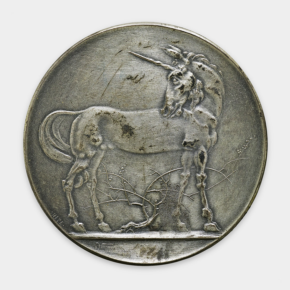 A unicorn is depicted in raised relief at the center of this circular silver medal. The unicorn is shown in profile, facing left, with a prominent horn extending from its forehead. Its body features a flowing mane and tail, and all four hooves are on the ground, with its legs slightly bent. The medal has a smooth, polished metallic surface. The background is plain, with thin tree branches visible behind the unicorn’s legs at the bottom. The edges of the medal are slightly raised, creating a defined border around the central image. The overall color of the medal is a uniform metallic silver.