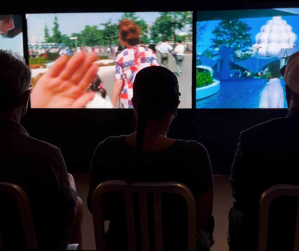 Black-and-white photograph of a man seated at a drafting table, holding a pencil, with large architectural drawings mounted on the wall behind him.