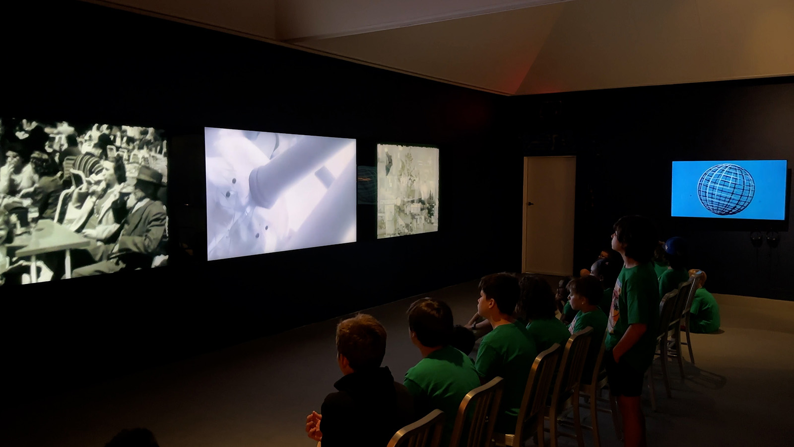 Young visitors seated in a dark gallery watching multiple illuminated screens on a black wall