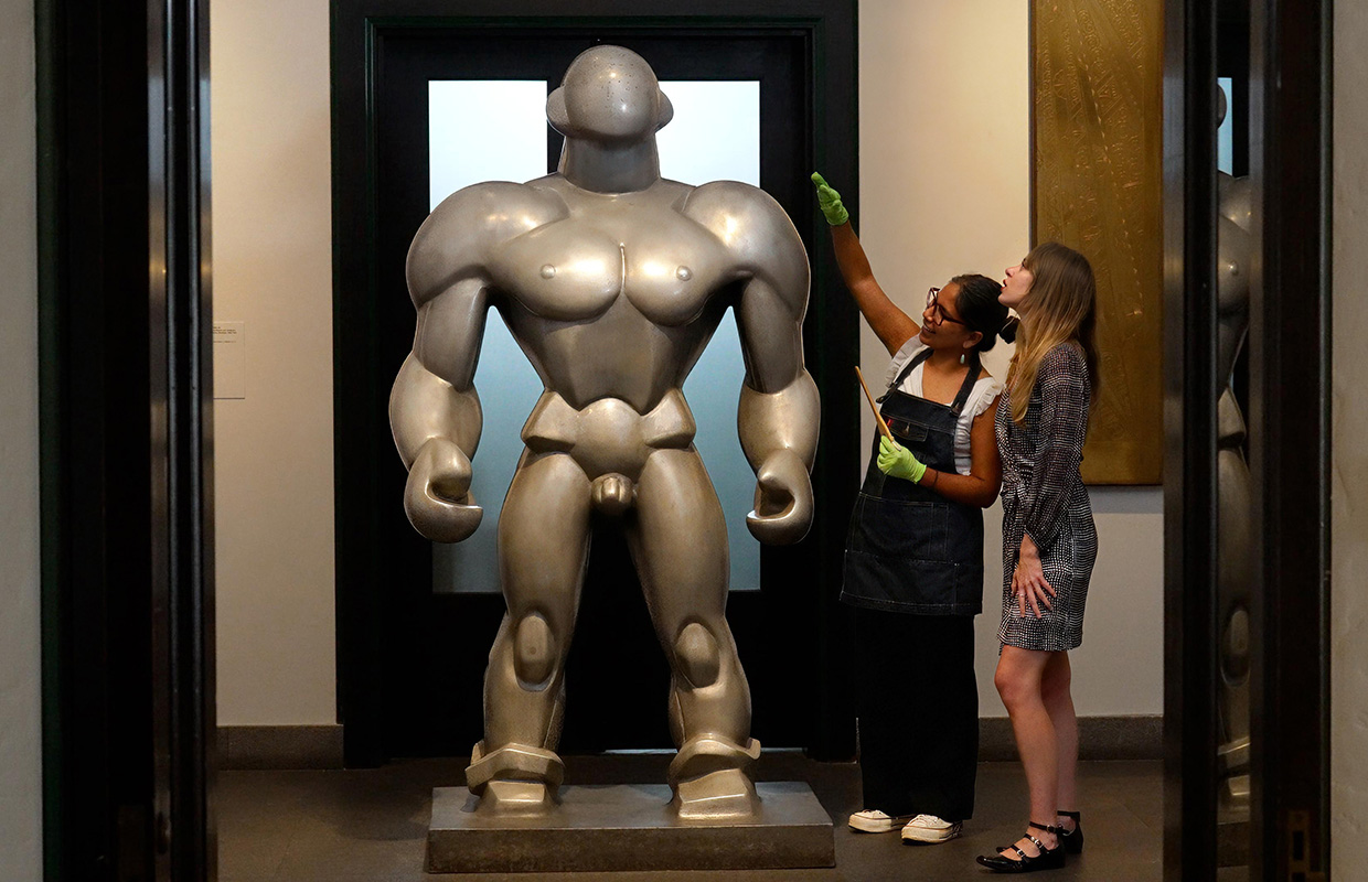 Two people examine a large silver metallic sculpture of a muscular, faceless figure in a hallway