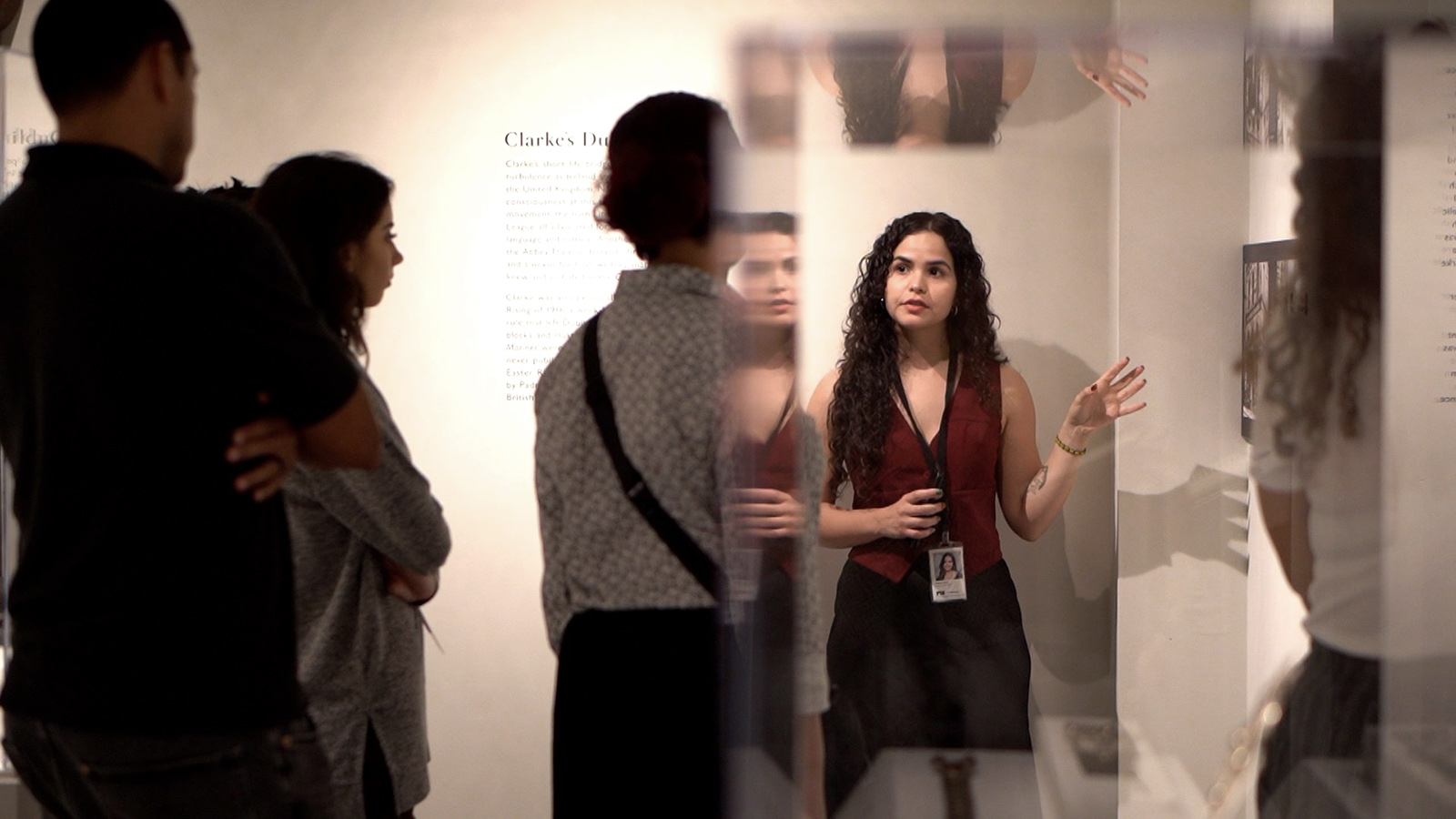 A woman leads a group tour in a museum gallery, gesturing toward an artwork on the wall while visitors listen