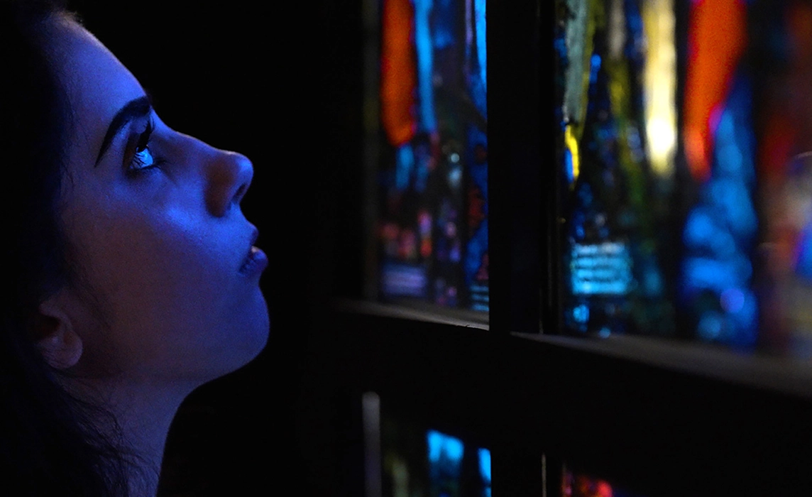 A detailed view of a woman looking closely at a multicolored stained glass window pane, with light reflecting on her face