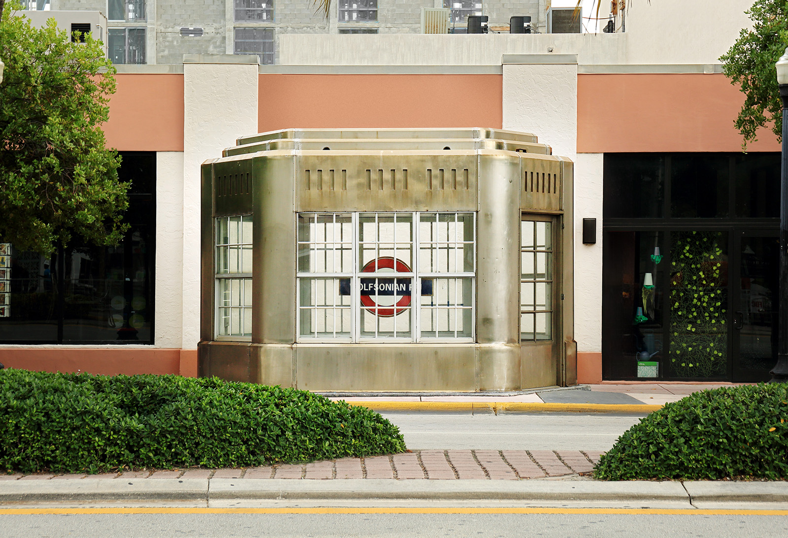 Street view of a building with a distinctive architectural feature in the center, which appears to be a rounded entrance with decorative metalwork. The building has a modern design with large windows and a flat roof. On either side of the central entrance, there are storefronts with large glass windows displaying various items. The storefront on the left has a sign that is not clearly visible, while the one on the right has signs that read "Design Store" and "Coffee Bar." There are trees and shrubs in front of the building, and a sidewalk runs along the street in front of it.