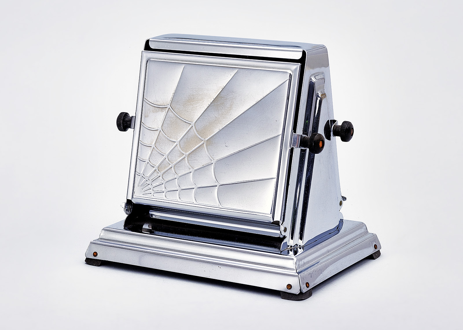 A chrome vintage toaster with a geometric, spiderweb-like design embossed on the side panel, black knobs on each side, and a rectangular base with rounded corners.