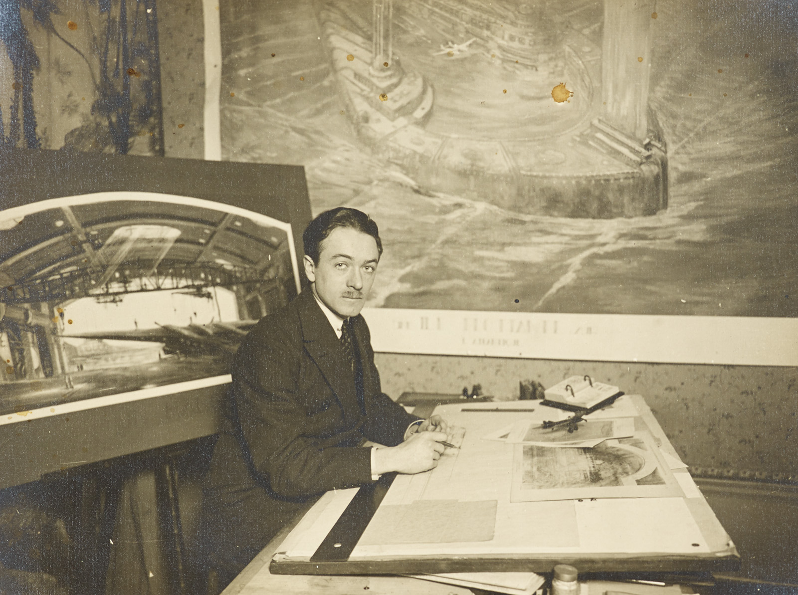 Man seated at a drafting table with architectural drawings and large renderings behind him