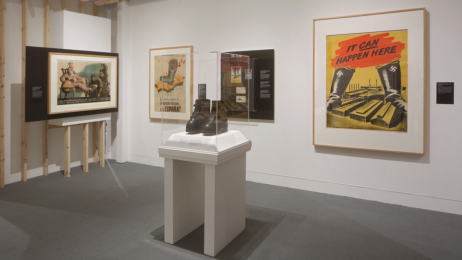 Gallery view with framed posters, a pair of black boots in a central case, and a large poster reading “It Can Happen Here”