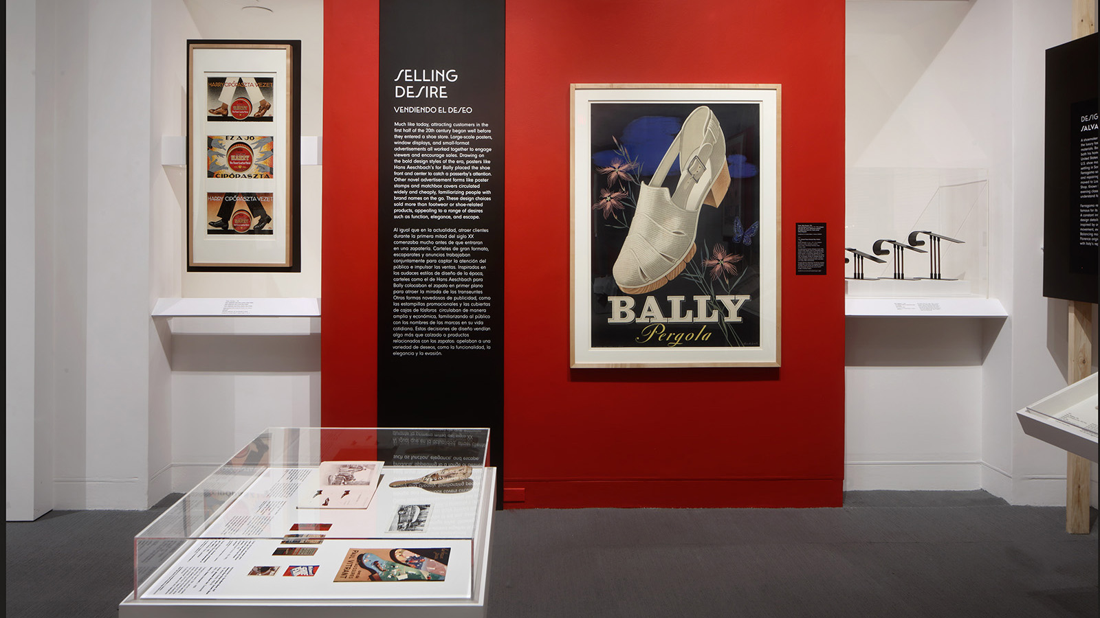 Gallery view with a red wall, a “Selling Desire” text panel, framed shoe advertisements, display cases, and three black shoe forms