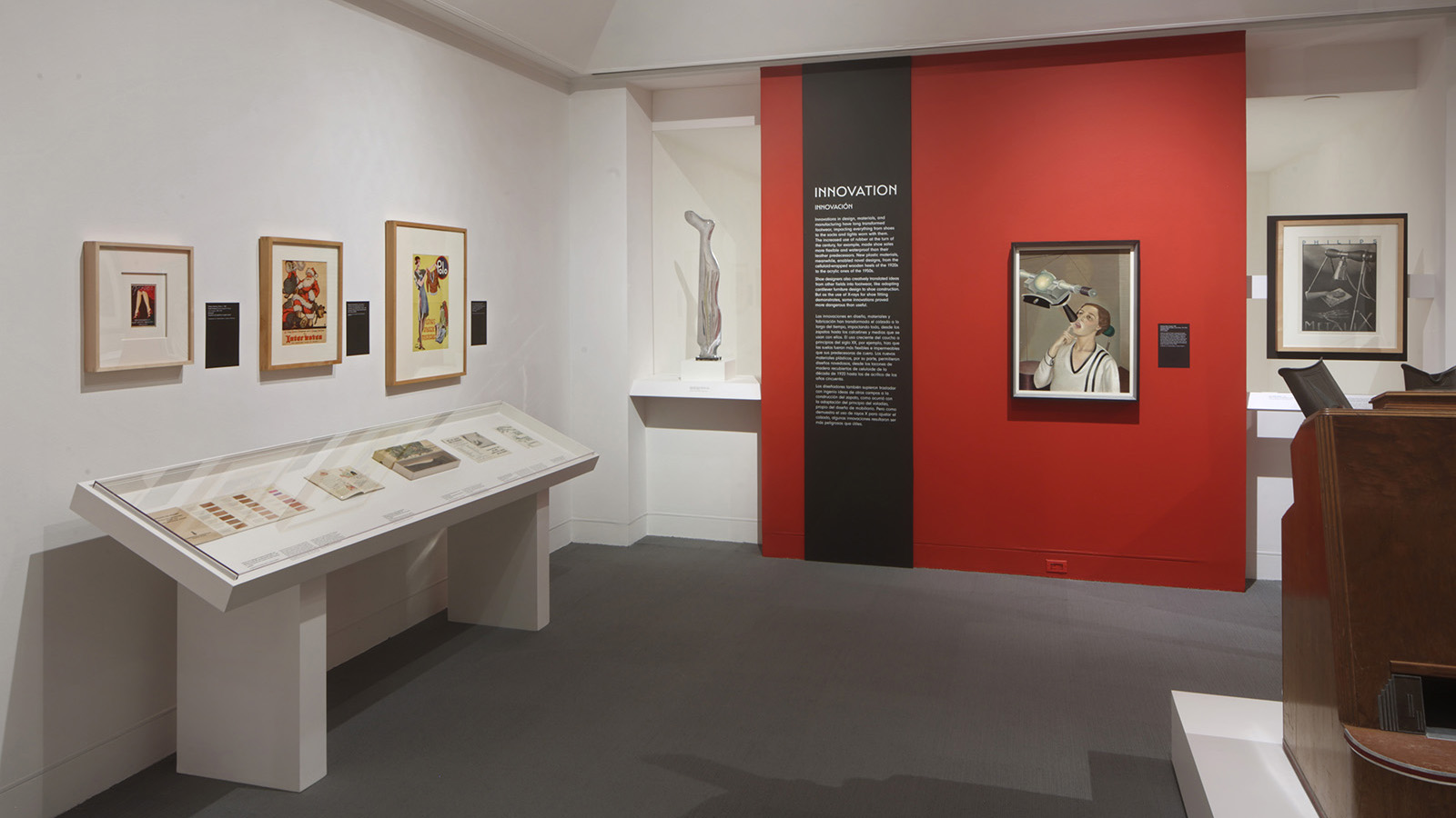 Gallery view with an “Innovation” text panel on a red wall, framed works, display cases, and a large wooden object at right