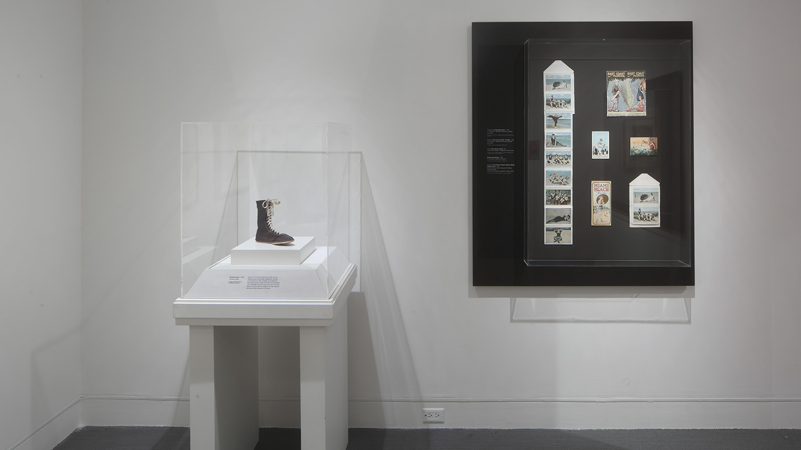 Black lace-up boot in a clear display case beside a framed arrangement of illustrated paper materials
