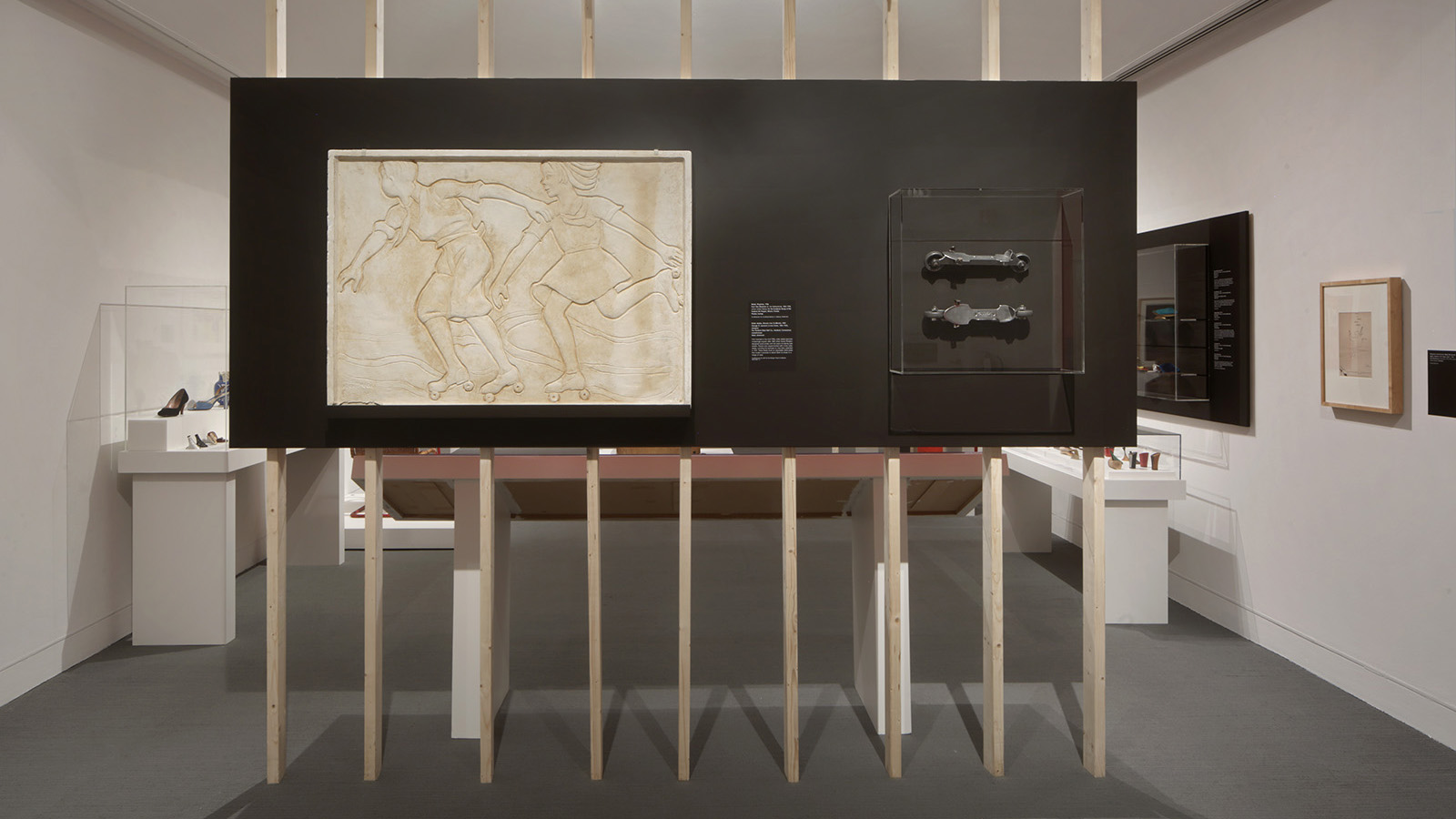 Freestanding black display wall with a relief of roller skaters and two metal objects in a clear case