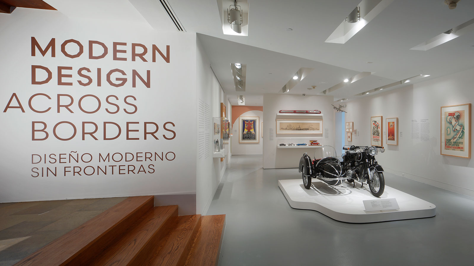 View of gallery with large wall text reading “Modern Design Across Borders / Diseño Moderno Sin Fronteras,” framed posters, display cases, and a motorcycle with sidecar on a central platform.