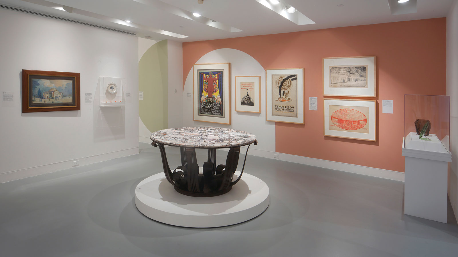 View of gallery with framed posters and drawings on white and peach-colored walls, a round marble-top table displayed on a circular platform, and a small sculptural object in a case to the right.