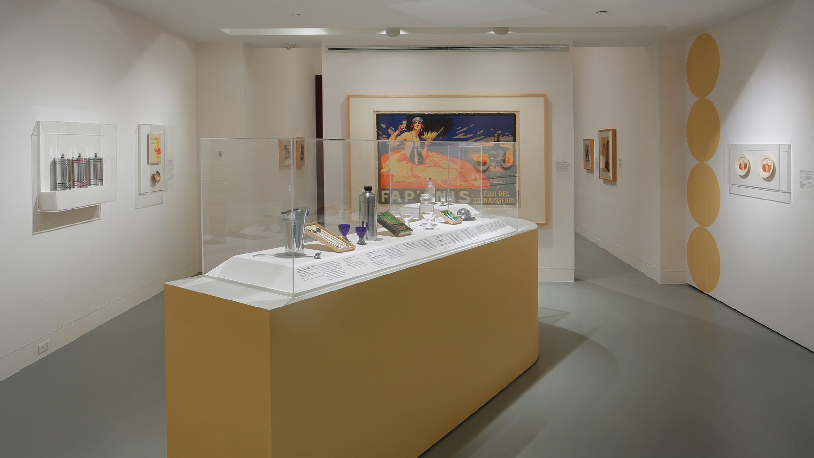View of gallery with a central display case holding small objects, surrounded by framed posters and works on paper on white walls.