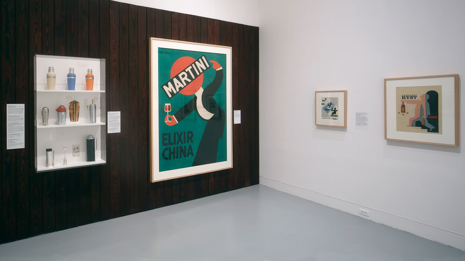 A wood-paneled gallery wall displays a large “Martini Elixir China” poster beside a case of cocktail shakers and barware, with two smaller framed graphic artworks on the adjacent white wall.