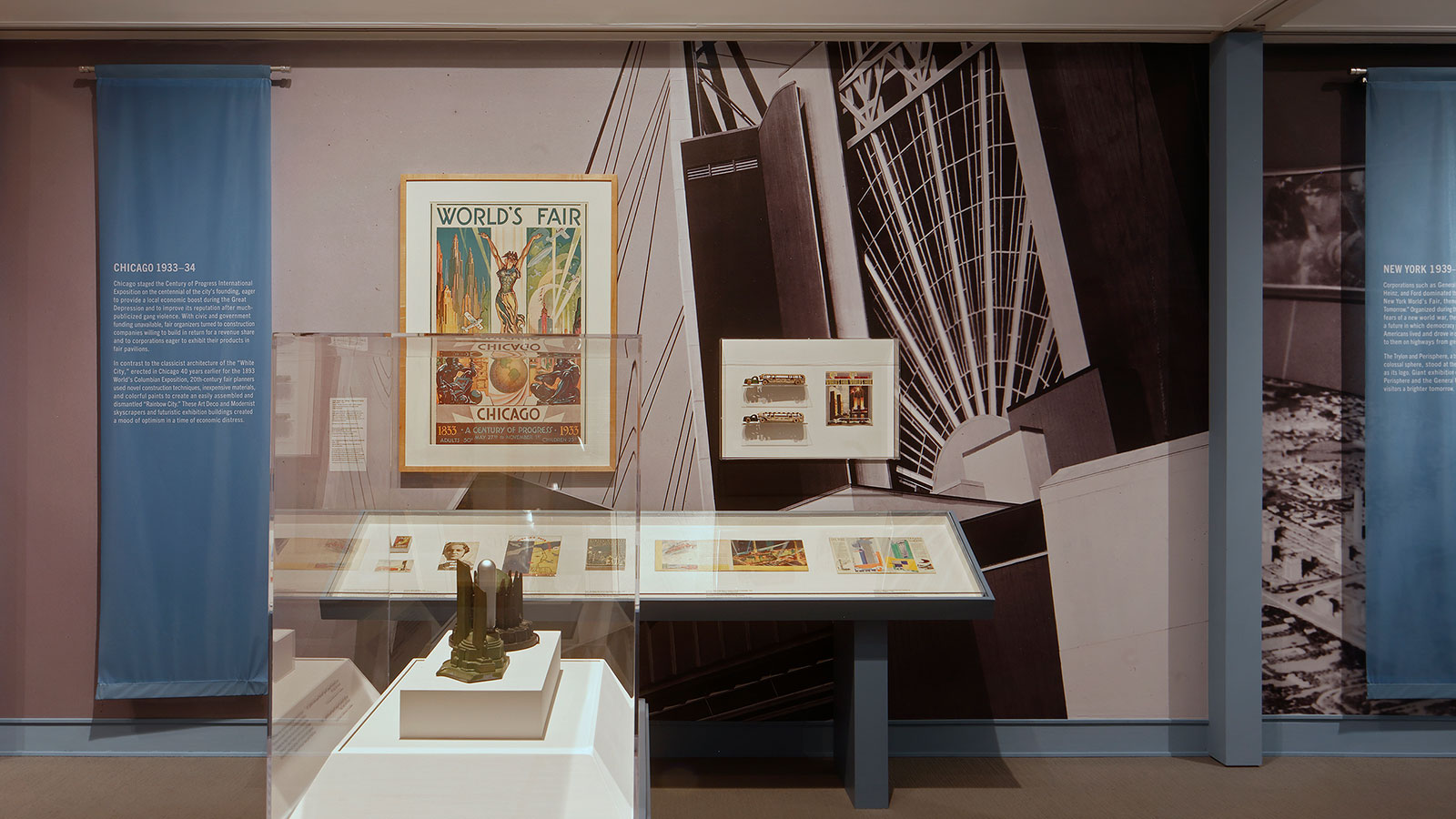 A gallery interior features a 1933 Chicago World’s Fair poster hung on a wall decorated with a large black-and-white architectural image. In front of the wall are historical ephemera and objects displayed in cases.