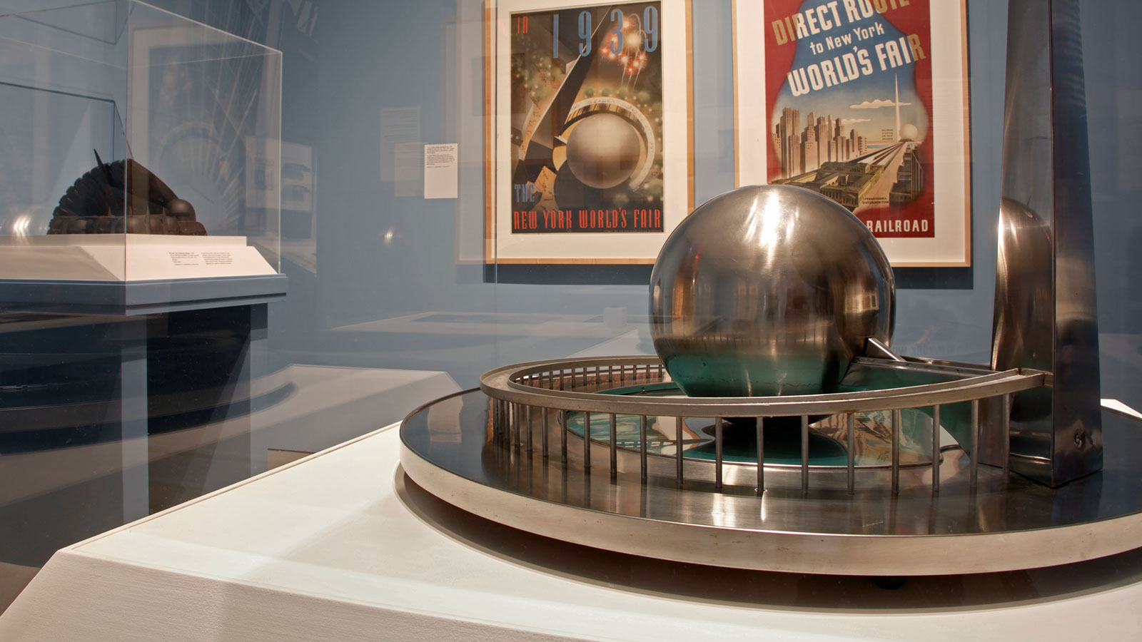 A metallic sculpture with a reflective sphere and tower on a circular base is displayed in a glass case in a gallery interior, with framed 1939 New York World’s Fair posters and other objects visible in the background.