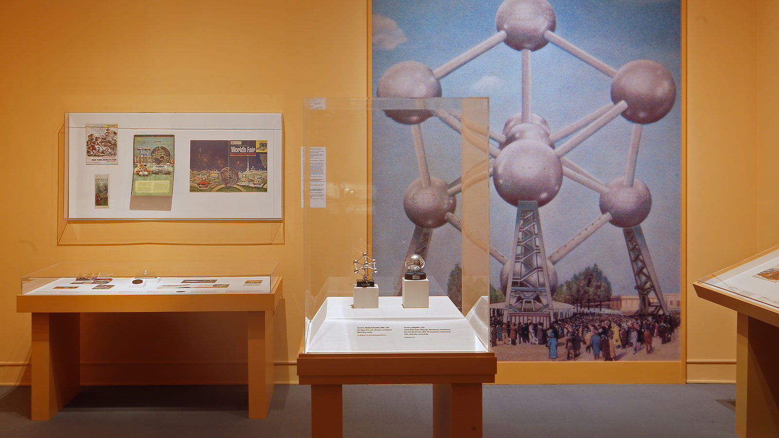 A gallery interior includes display cases of historical artifacts and two souvenirs installed in front of a yellow wall decorated with a large reproduction image of the Atomium structure from Expo 58 in Brussels.