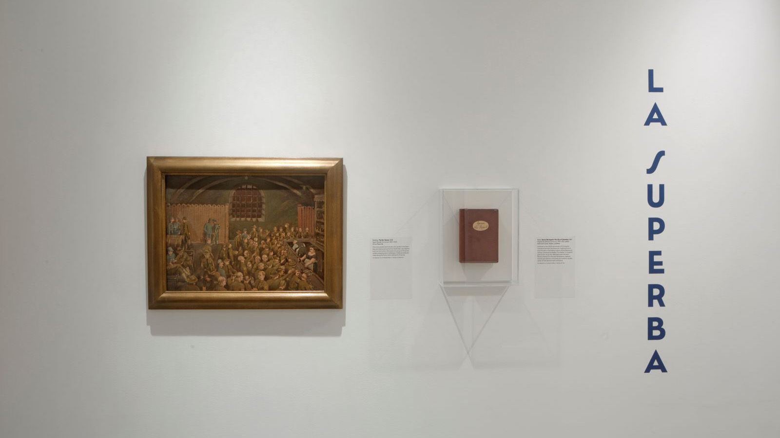 Framed painting and red book mounted on a white wall with the blue text “LA SUPERBA” arranged vertically to the right.